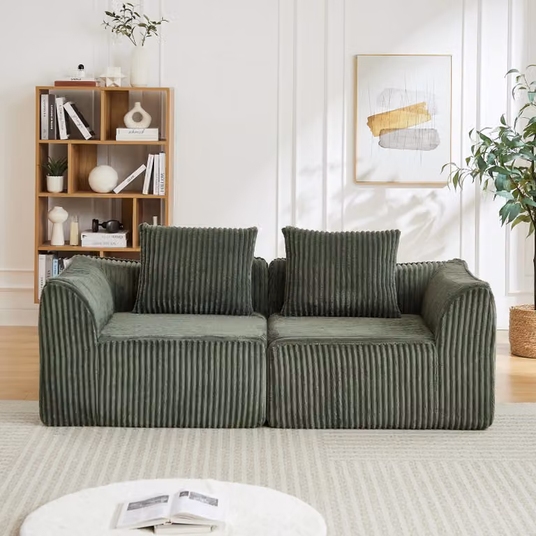 Minimalistyczna sofa tapicerowana tkaniną corduroy, o zaokrąglonych kształtach, z delikatnie opadającym oparciem, z dwoma poduszkami